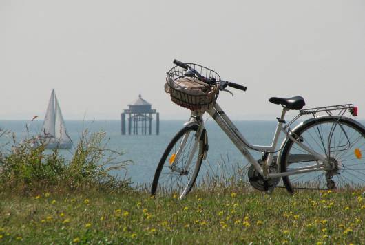 Vélo au premier plan avec en fond la mer, un voilier et le phare du bout du monde près de La Rochelle